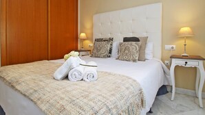 Uma cama bem arrumada com cabeceira branca, mesa de cabeceira com abajur e guarda-roupa de madeira.