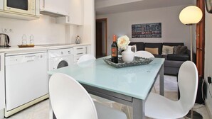 Cozinha moderna com eletrodomésticos brancos, uma mesa de jantar com cadeiras e uma sala de estar com sofá e abajur.