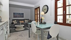 Uma sala de estar moderna com um sofá, uma mesa de jantar com cadeiras, um relógio e uma janela com vista para prédios.