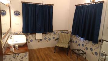 Een badkamer met een wastafel, spiegel, blauwe gordijnen, bloemenbehang, een stoel en een klein tafeltje.