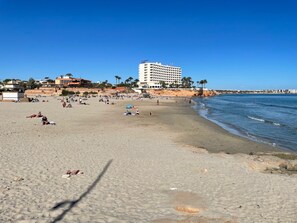 Una playa con un hotel grande al fondo, orillas arenosas y cielos azules despejados.