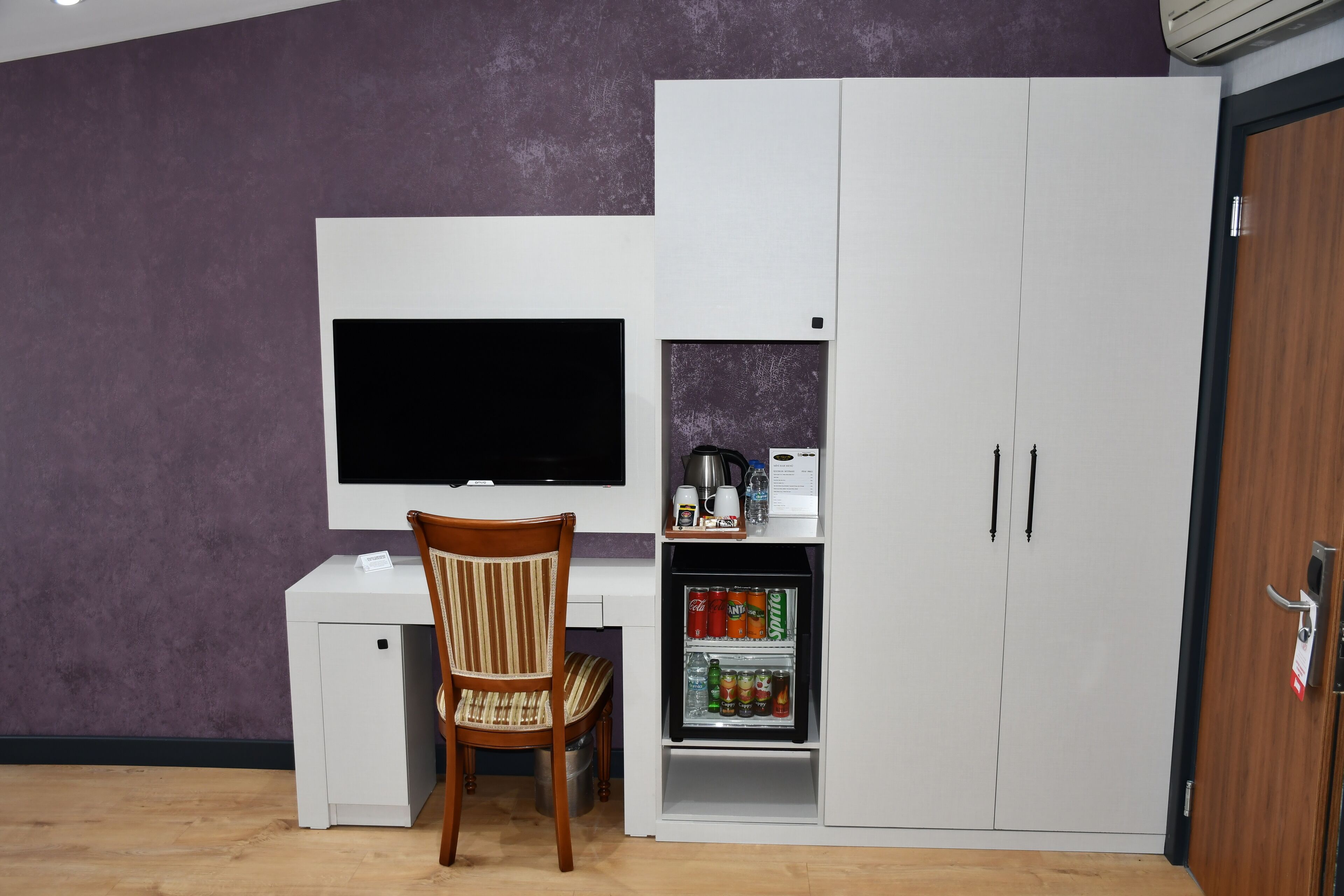 Habitació senzilla amb escriptori blanc, televisió, nevera petita amb begudes i un armari.