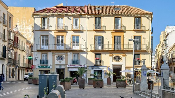 A multi-story building with a tiled roof, multiple balconies, and storefronts including a pharmacy and a restaurant. - Historic Málaga Apt: Quiet Retreat w/ Courtyard, 4-min Walk to Larios (Málaga)