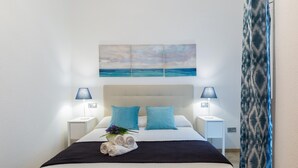 A neatly made bed with a white headboard, two pillows, and a rolled towel with flowers. There are two bedside lamps, a wall-mounted painting of a beach, and a patterned curtain. - Historic Málaga Apt: Quiet Retreat w/ Courtyard, 4-min Walk to Larios (Málaga)