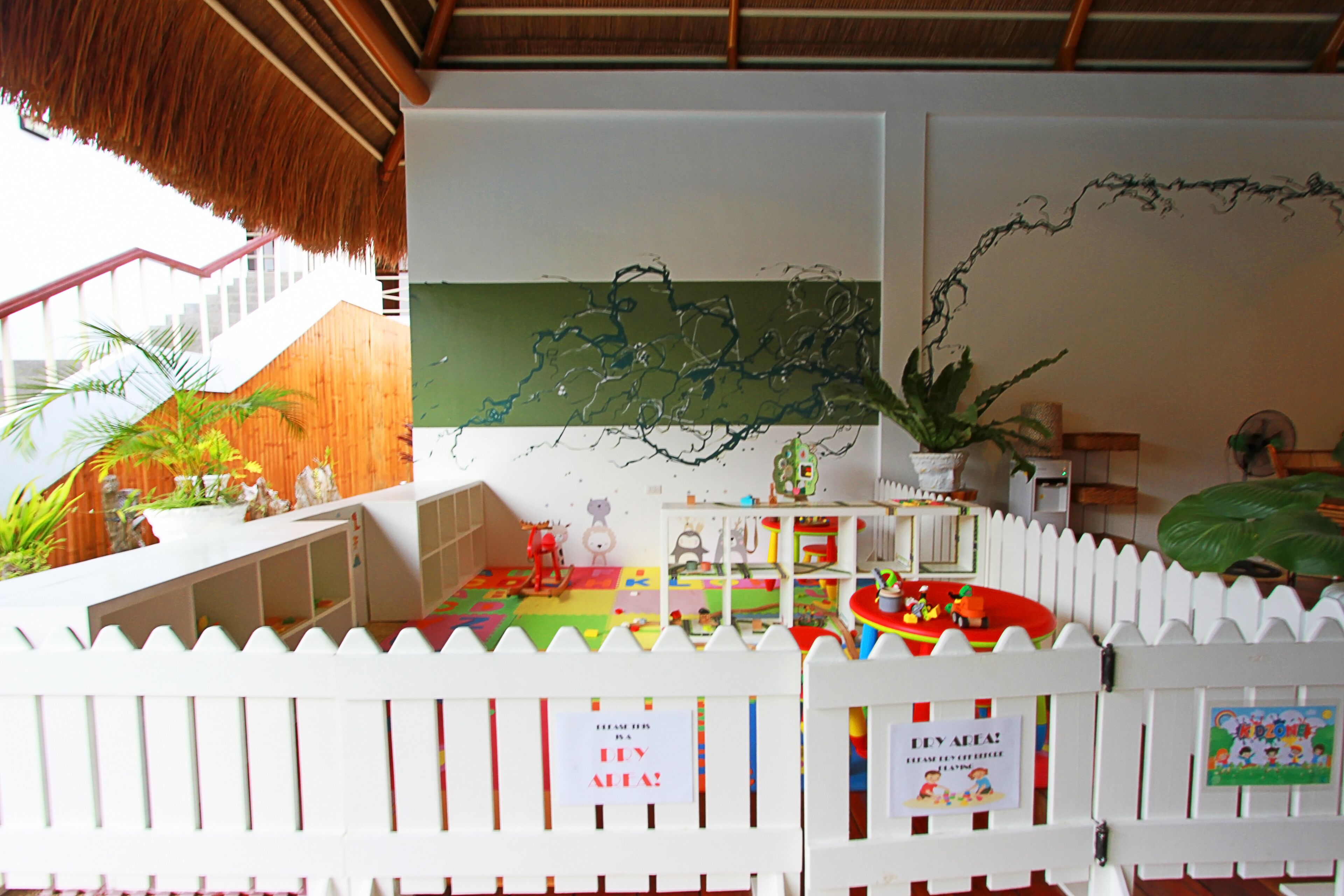 Un área de juegos infantiles con cerca de piquetes blancos, techo de paja y varios juguetes y estructuras recreativas.