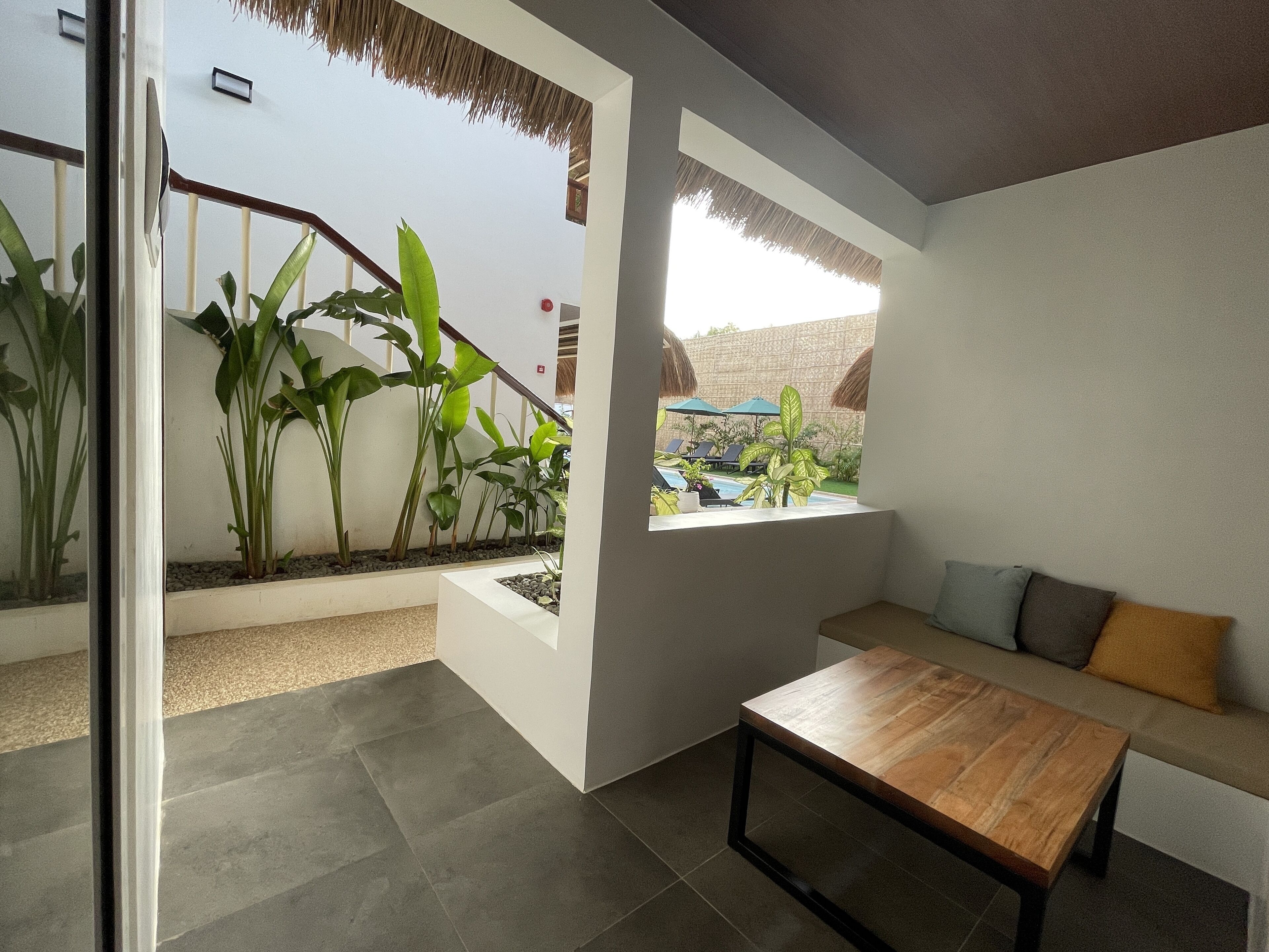 A modern living room with a wooden coffee table, a sofa with cushions, and a large window overlooking a pool area with lounge chairs and umbrellas.