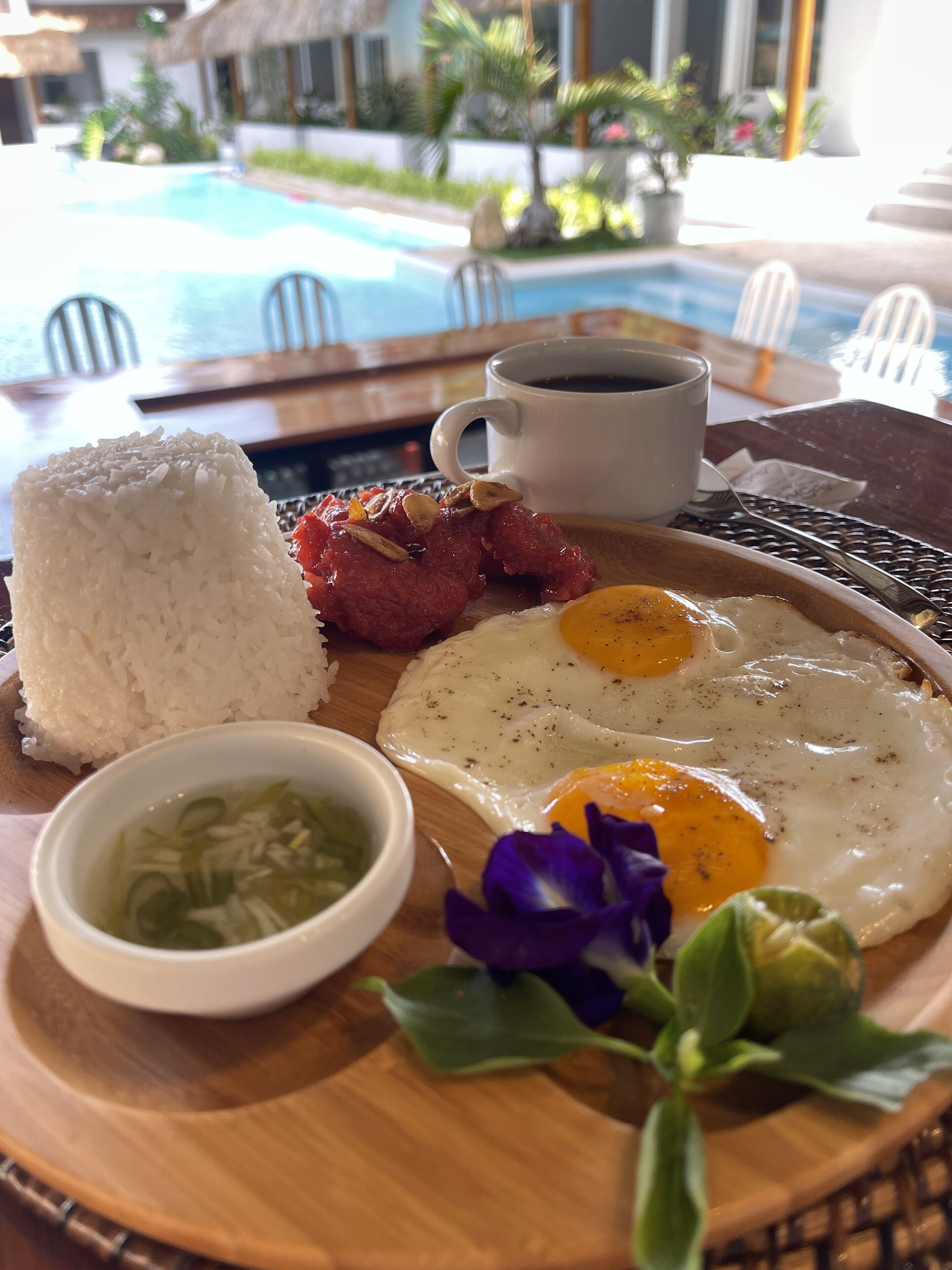 A breakfast tray with rice, eggs, and a small bowl of soup, set on a wooden table with a view of a pool and chairs in the background.