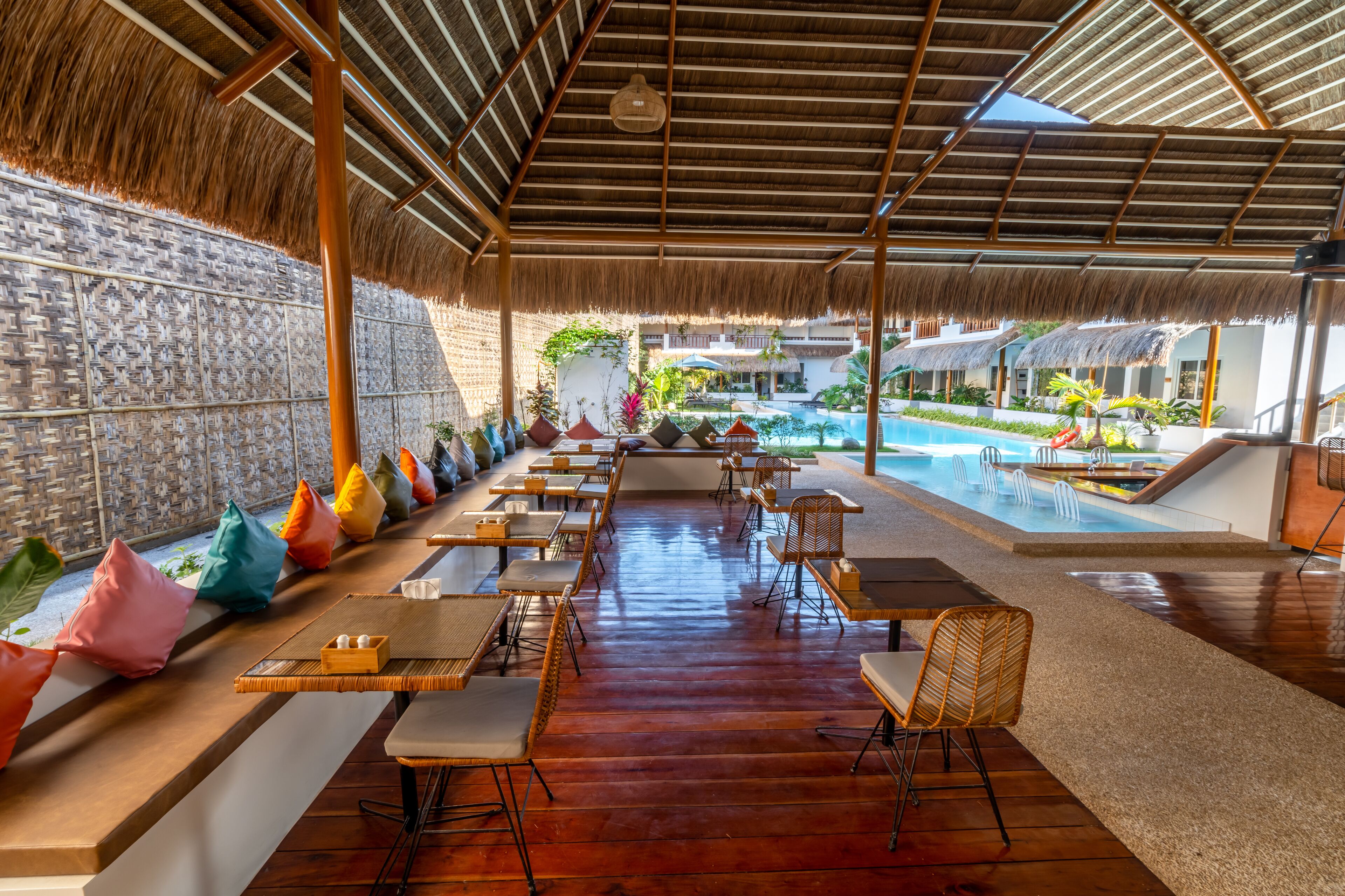 Un área de comedor al aire libre con mesas y sillas de madera, un techo de paja y una piscina al fondo.