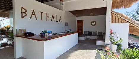 Un espace d’accueil avec un comptoir portant l’inscription « BATHALA », un coin salon avec des coussins et une structure à toit de chaume.