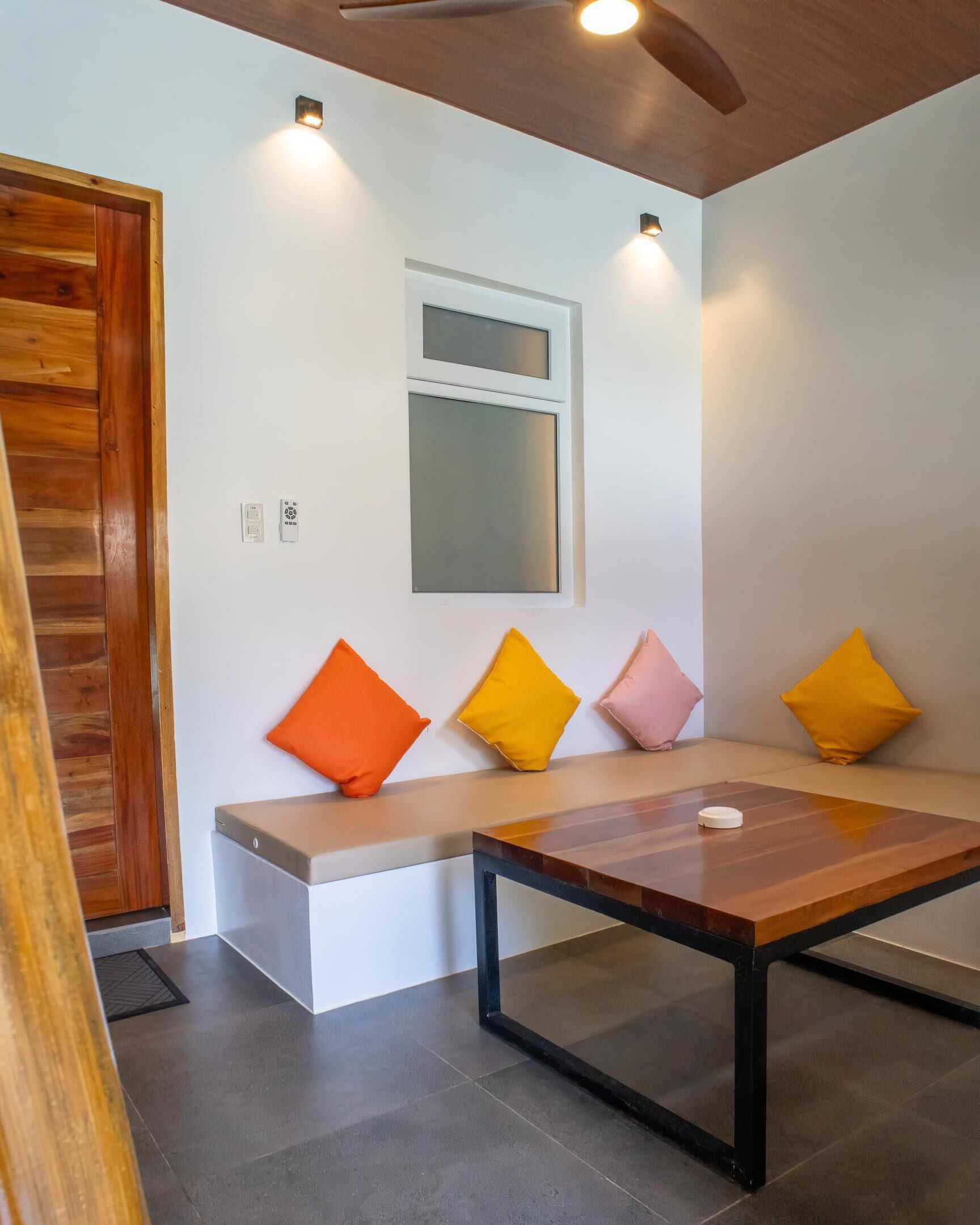 Une pièce moderne avec une porte en bois, un banc agrémenté de coussins colorés, une table et un éclairage encastré au plafond.