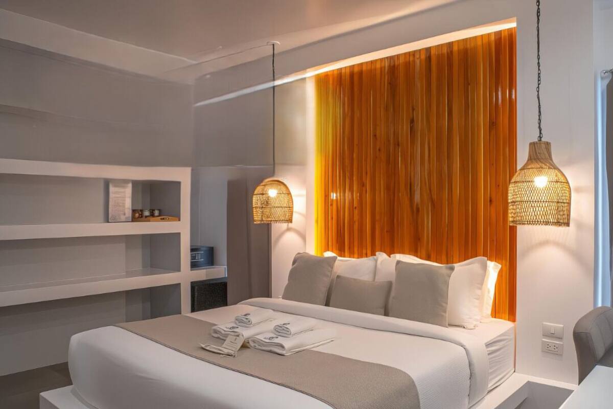 Un dormitorio moderno con una cama grande, ropa de cama blanca y una pared de acento de madera. Hay dos lámparas colgantes sobre la cama y una estantería a la izquierda.