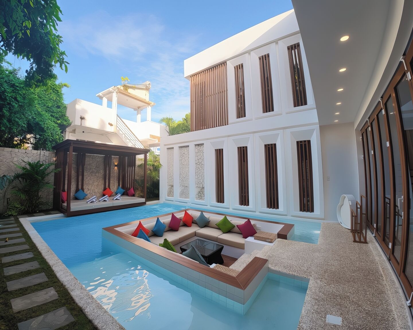 A modern pool area with a lounge chair and a covered seating area, surrounded by a stone wall and greenery.