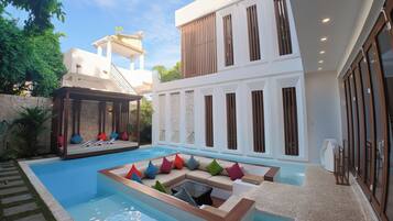 A modern pool area with a lounge chair and a covered seating area, surrounded by a stone wall and greenery.