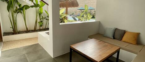 A modern living room with a wooden coffee table, a sofa with cushions, and a large window overlooking a pool area.