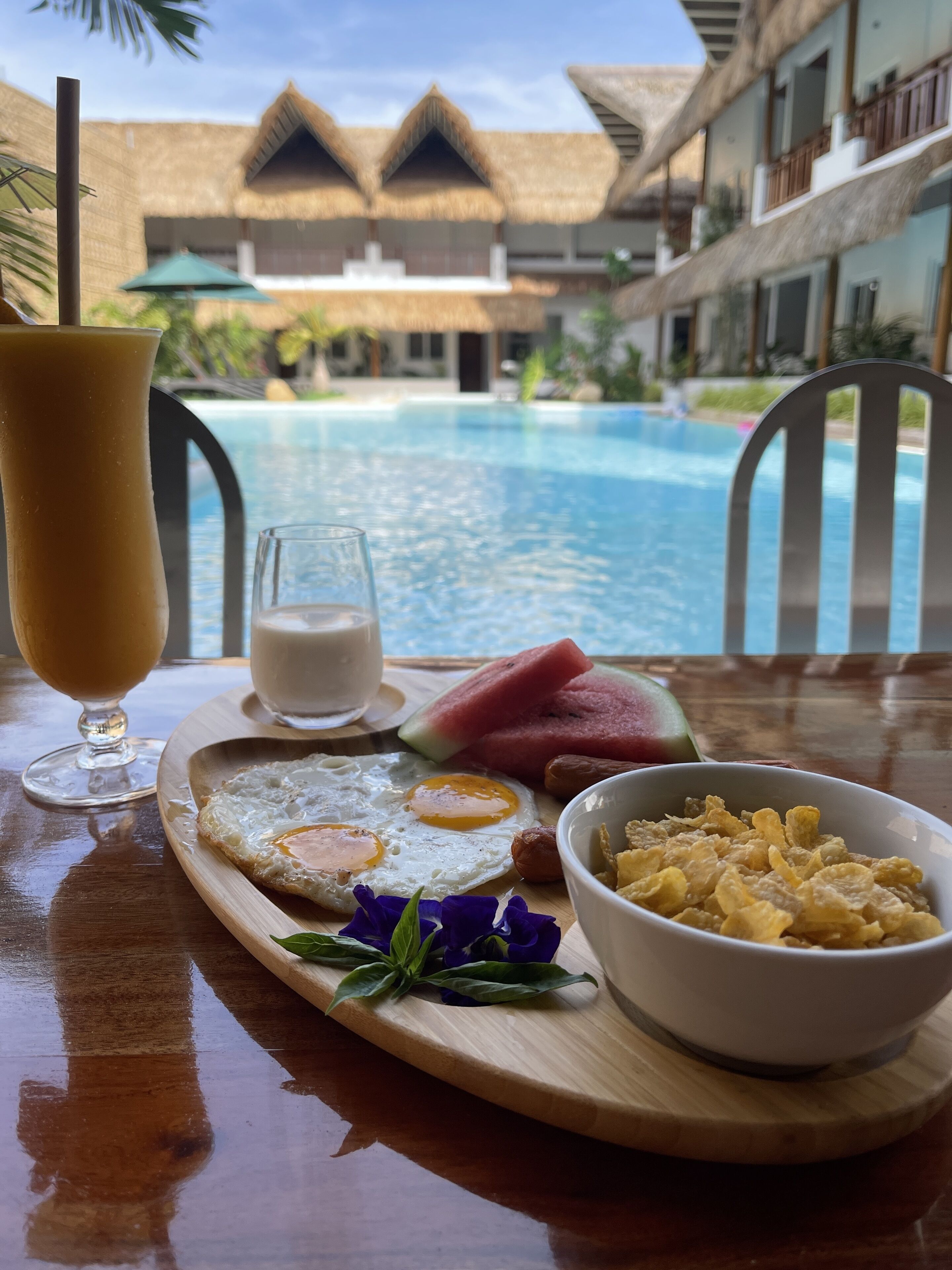 Bandeja de desayuno con huevos, tostadas, sandía y cereal, con una piscina y edificios de un complejo turístico al fondo.