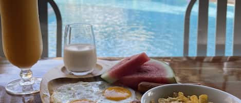 A breakfast tray with eggs, toast, watermelon, and cereal, with a pool and resort buildings in the background.
