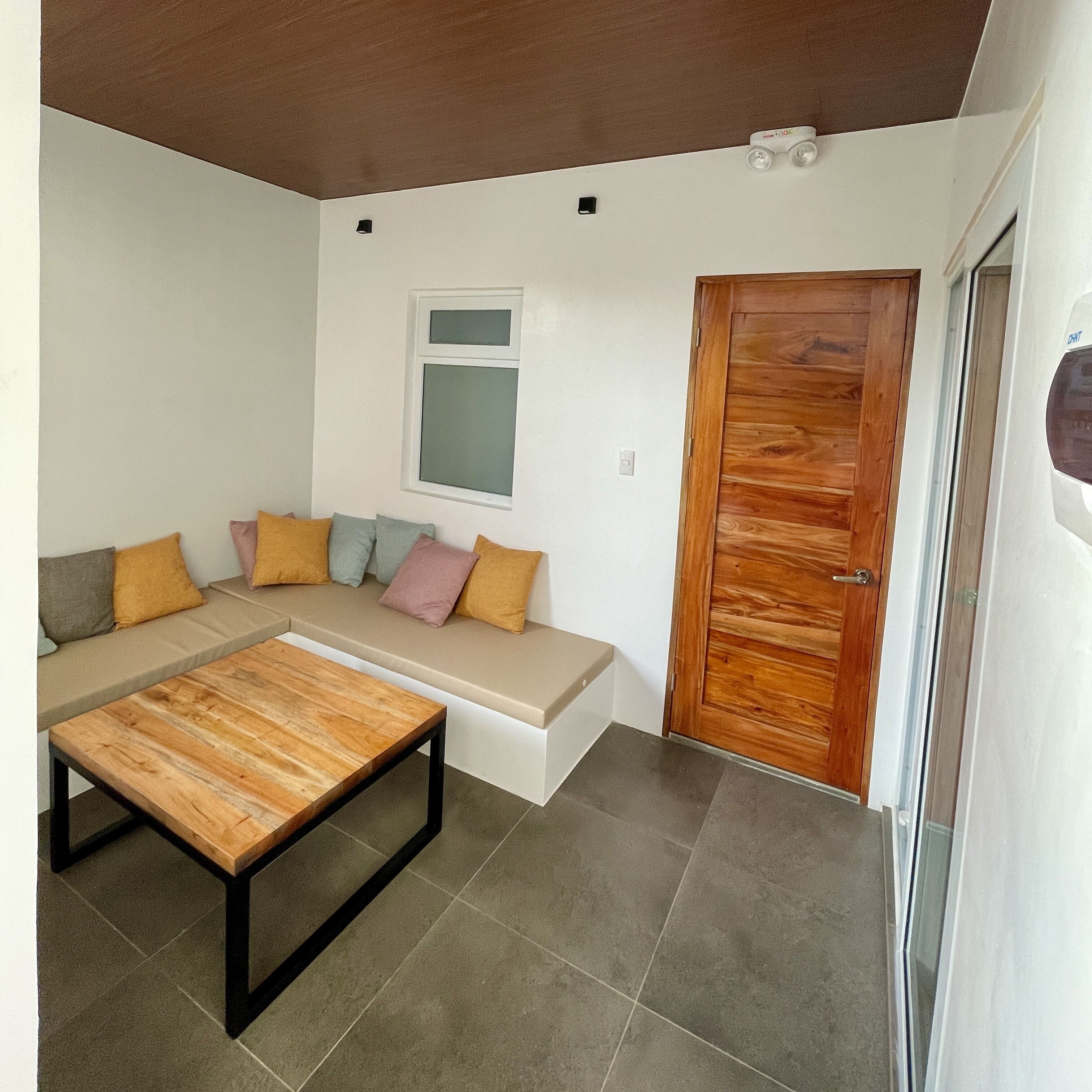 A living room with a wooden coffee table, a sofa with cushions, a wooden door, and a window.