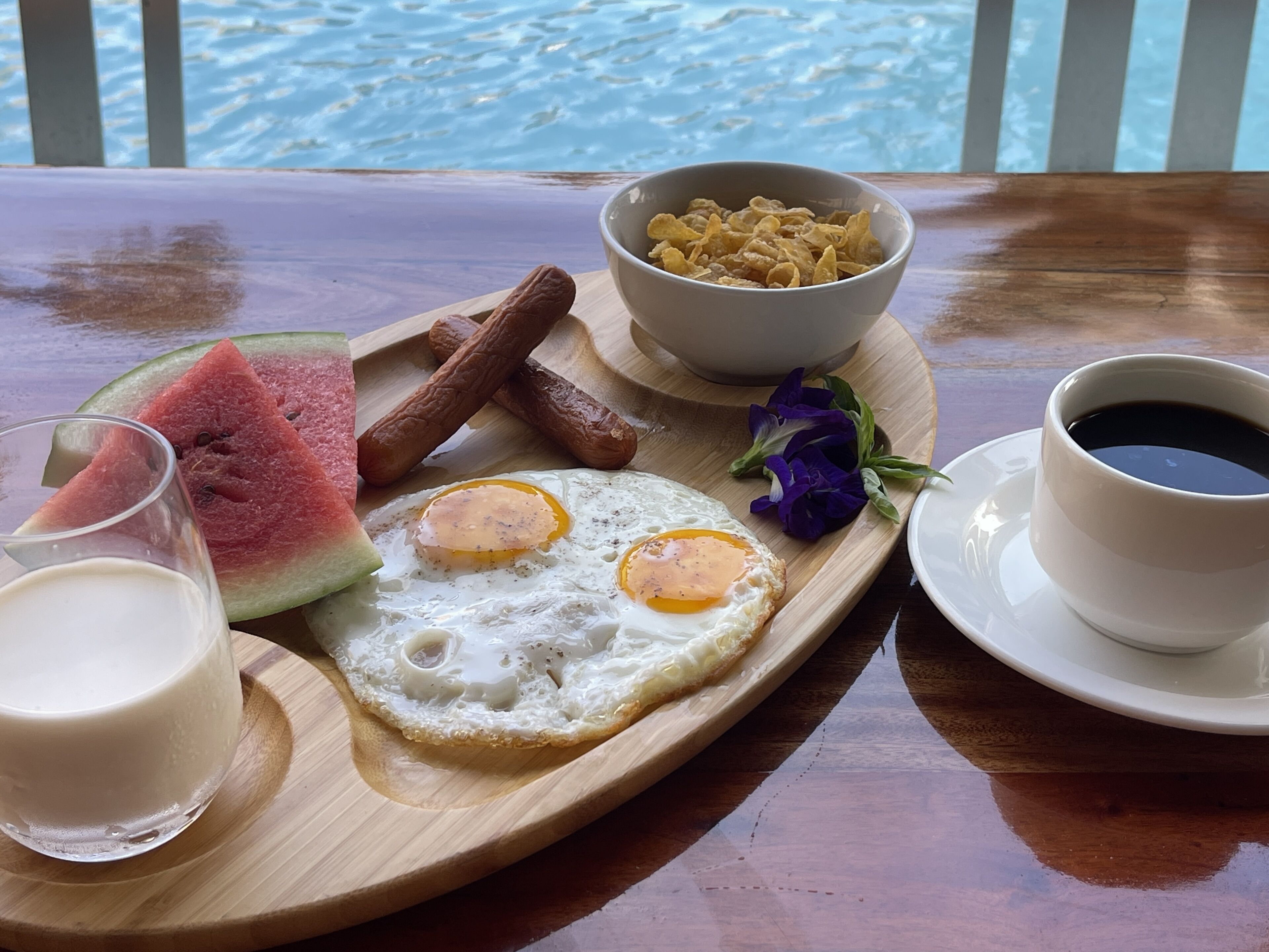 A breakfast tray with eggs, sausages, cereal, a slice of watermelon, a glass of milk, and a cup of coffee.