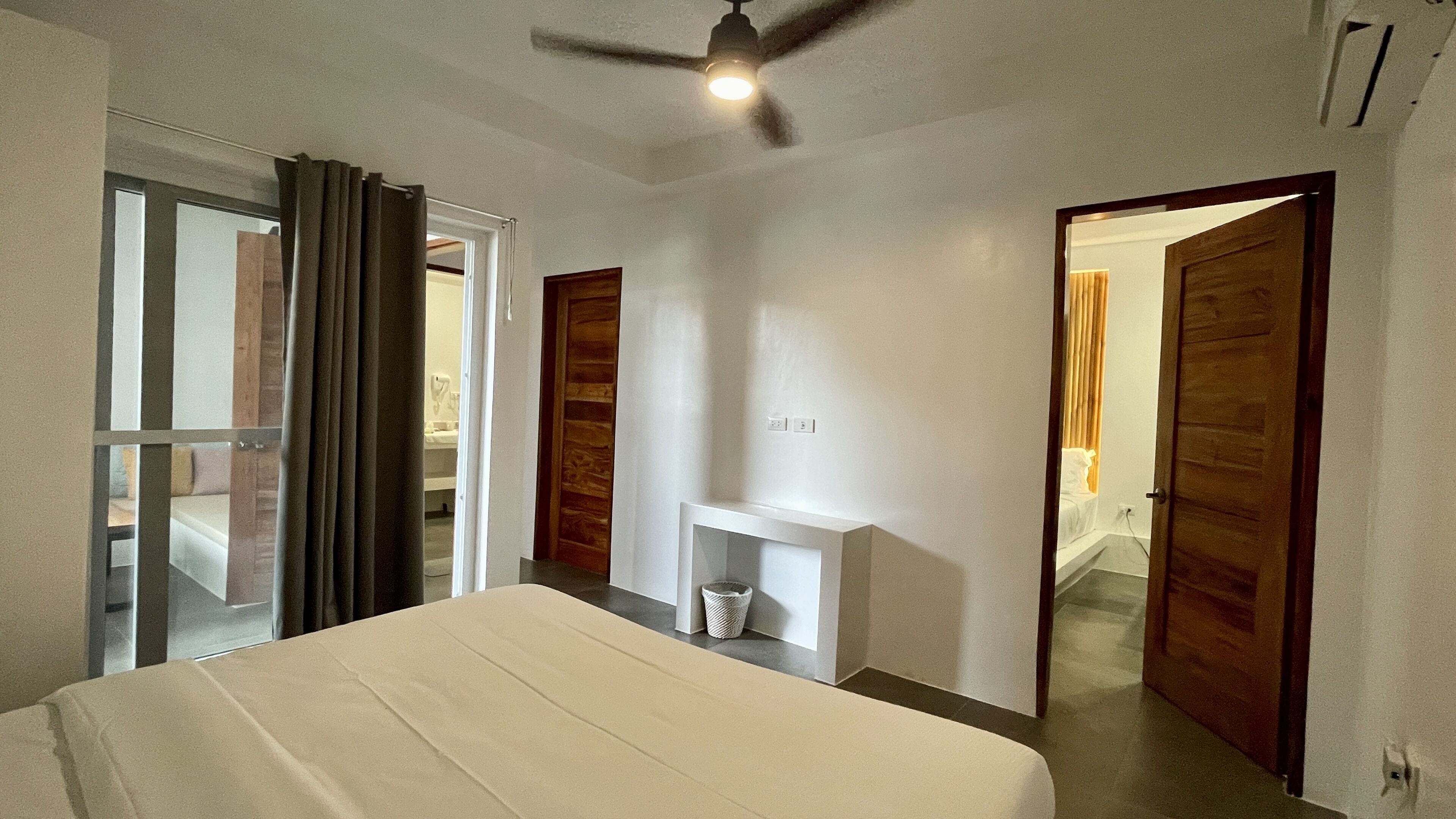A hotel room with a bed, a ceiling fan, a small white console table, and a wooden door.