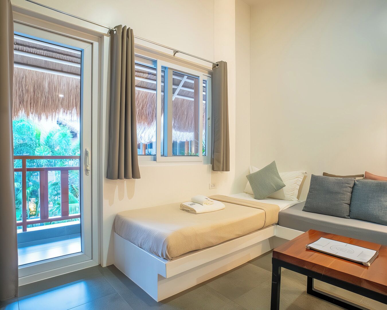 A compact room with a bed, a sofa, and a small table. There are windows with curtains, and a view of greenery outside.