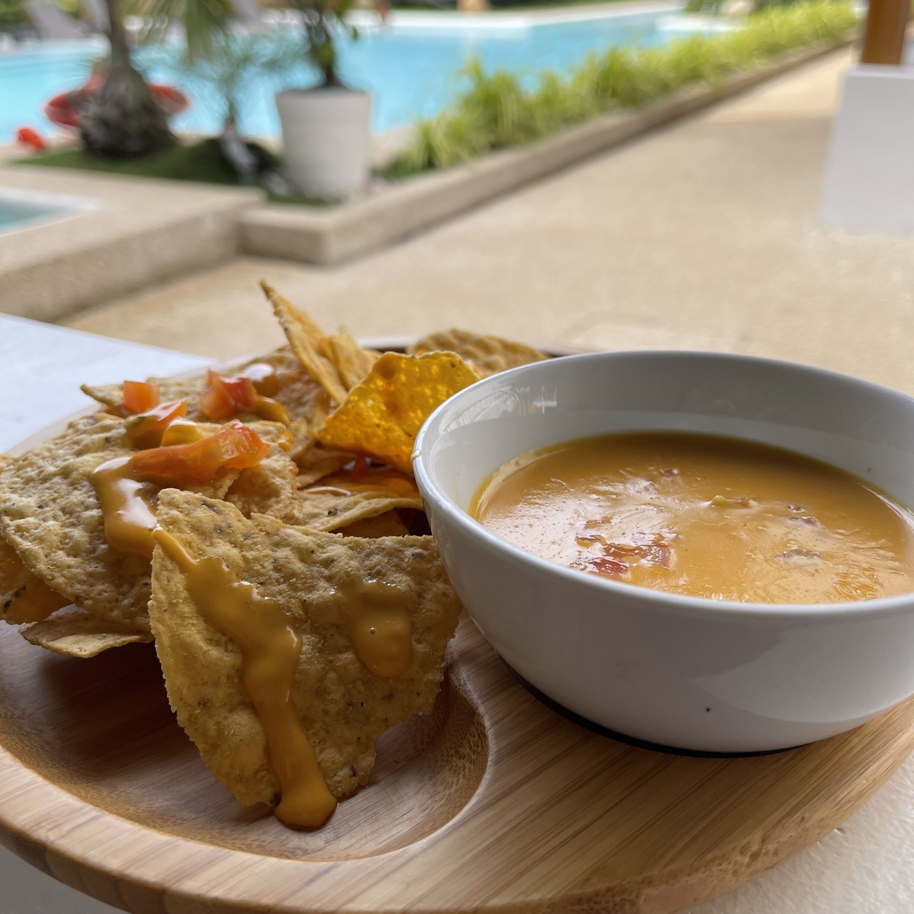 A wooden serving tray with nachos, a bowl of soup, and a pool in the background.