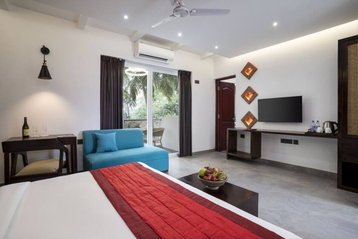 A modern hotel room with a bed, a blue sofa, a small table with a fruit bowl, a TV, and a desk with a lamp.