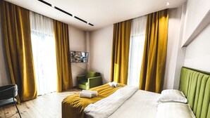 Habitación de hotel con cama, cabecera verde, cortinas amarillas y una silla verde.