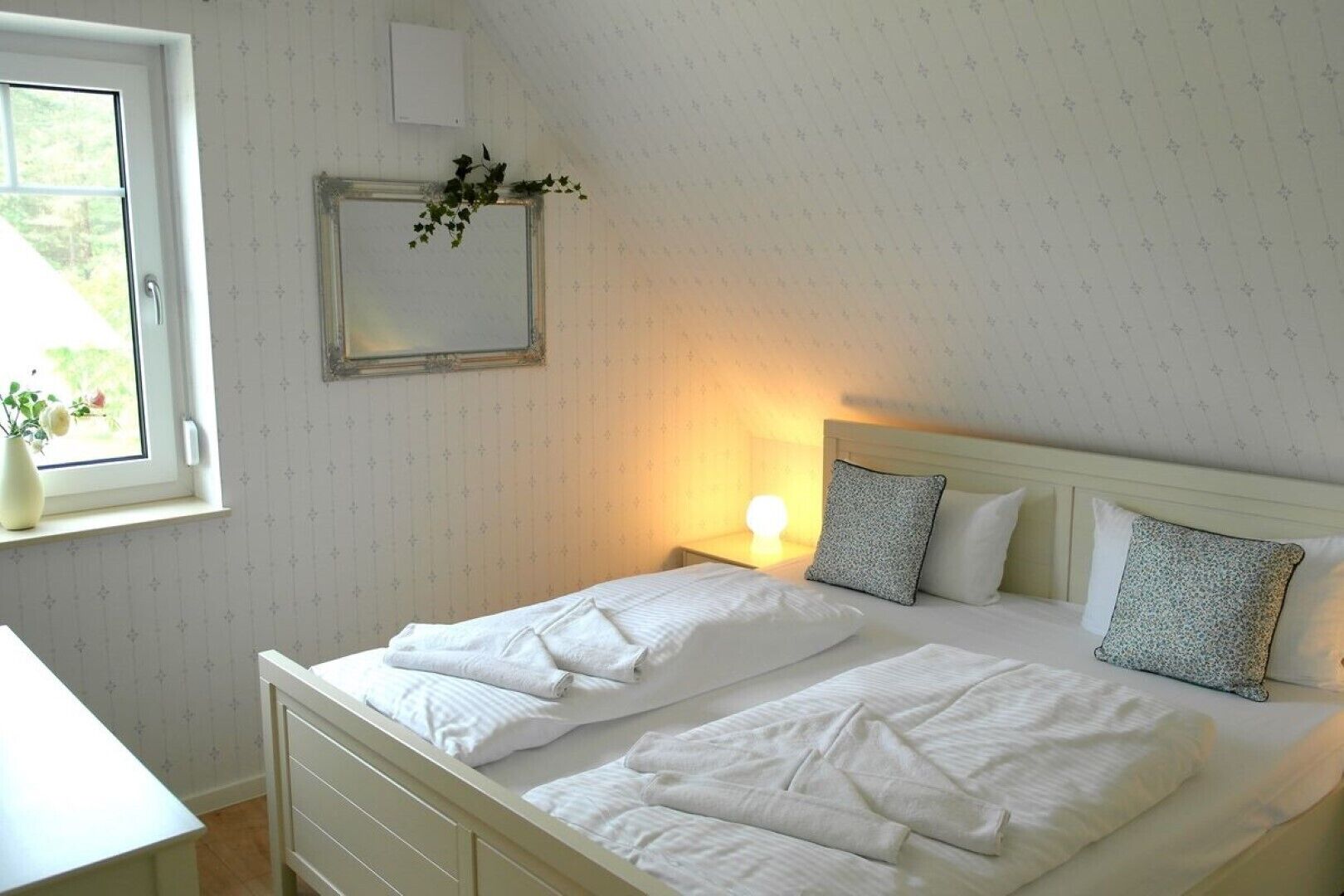 A neatly made bed with white linens and pillows, a bedside table with a lamp, and a window with a view of greenery.