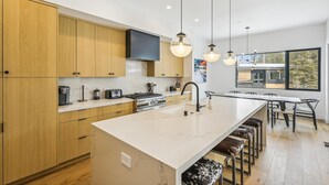 A modern kitchen with wooden cabinets, a large island, and a dining area with bar stools. - Diamond Run at Palisades by Tahoe Getaways - Luxury 4BR Townhome, Pet-Friendly, Shuttle & Hot Tub! (Olympic Valley)