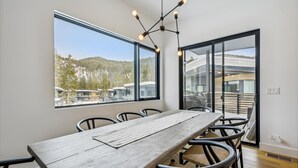 A modern dining area with a wooden table and chairs, large windows with a view of buildings and trees, and a ceiling light fixture. - Diamond Run at Palisades by Tahoe Getaways - Luxury 4BR Townhome, Pet-Friendly, Shuttle & Hot Tub! (Olympic Valley)