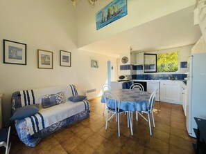 Un salon chaleureux avec un canapé et une table à manger, une cuisine avec des meubles blancs et des carreaux bleus, et un tableau représentant des voiliers accroché au mur.