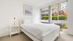 A bedroom with a bed, bedside table, and a window with a view of greenery and a building.