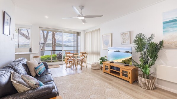 A modern living room with a leather sofa, a dining table, chairs, a television, and a large window offering a view of the sea.