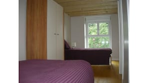 A bedroom with two beds, wooden ceiling beams, a window with a view of greenery, and a door leading to another room.