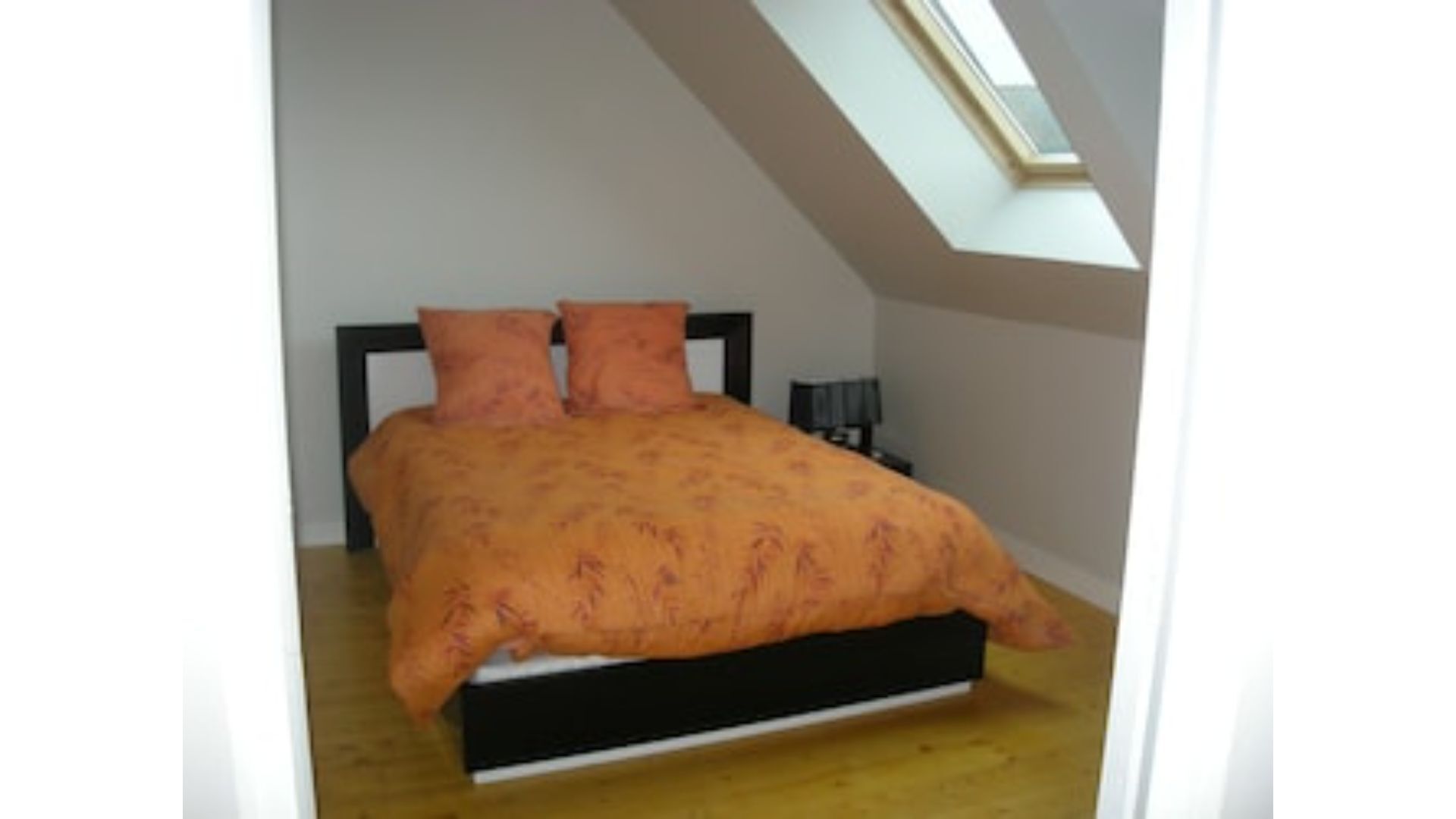 Un dormitorio con tragaluz, una cama con colcha naranja y una mesita de noche con lámpara.