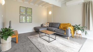 Una sala de estar moderna con un sofá gris, una mesa de centro de madera y una alfombra gris. Hay dos plantas en macetas, una sobre una mesita de madera y otra sobre un pequeño pedestal. La habitación tiene un techo blanco con vigas a la vista y una unidad de aire acondicionado montada en la pared.