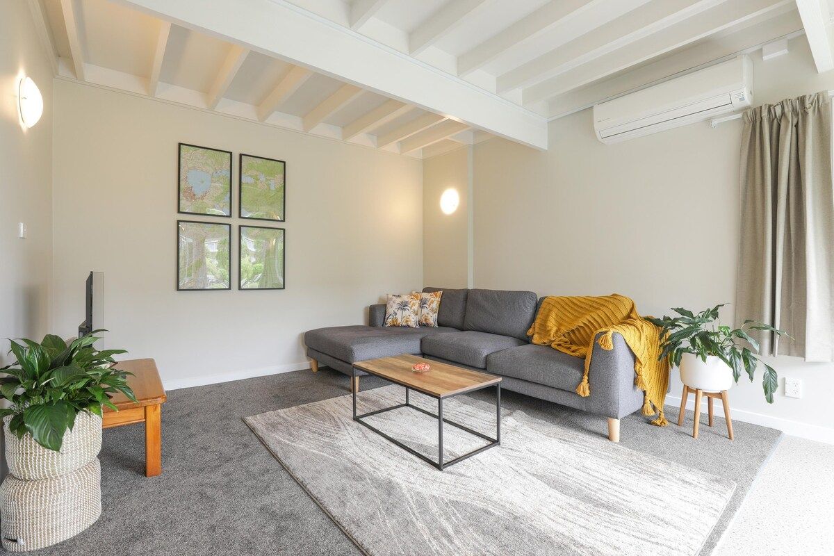 Un salon moderne avec un canapé gris, une table basse en bois et un tapis gris. On y trouve deux plantes en pot, l’une posée sur un tabouret en bois et l’autre sur le sol.