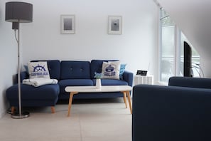 A modern living room with a blue sofa, a white coffee table, and a floor lamp. There are two framed pictures on the wall, and a laptop is on a small table.