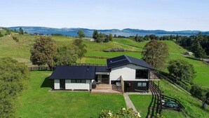 Une maison moderne à deux étages, dotée d’une grande terrasse, entourée de vastes prairies verdoyantes et avec un lac en arrière-plan.