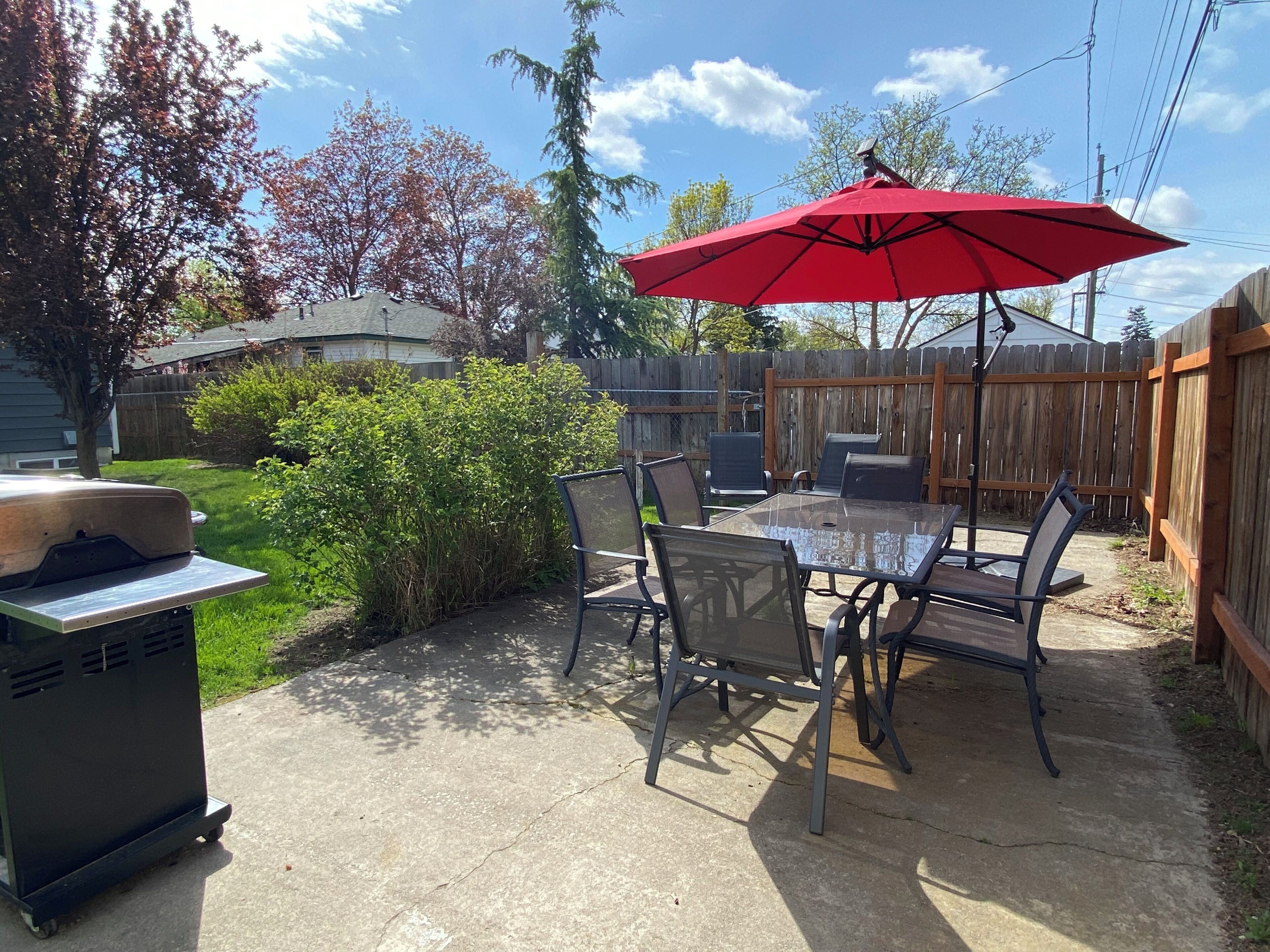 An outdoor patio with a table and chairs, a red umbrella, and a grill.