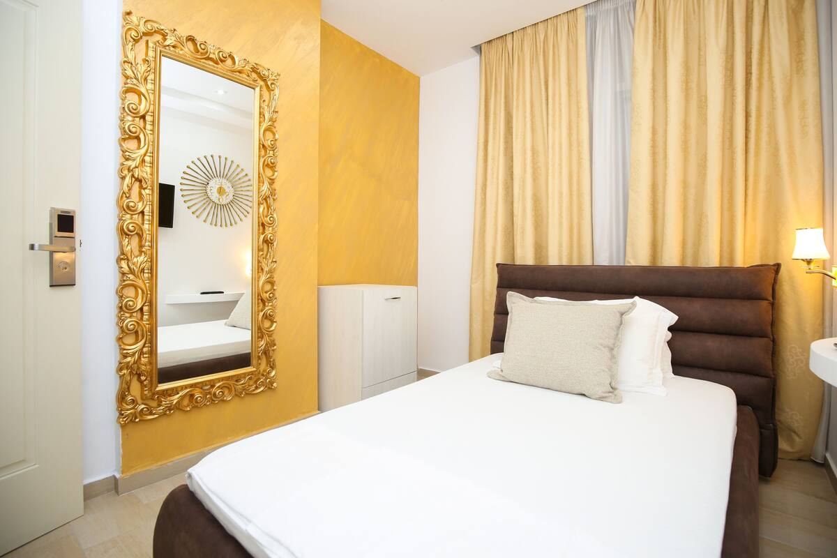 A hotel room with a large ornate mirror, a bed with two pillows, a bedside table with a lamp, and a small cabinet.