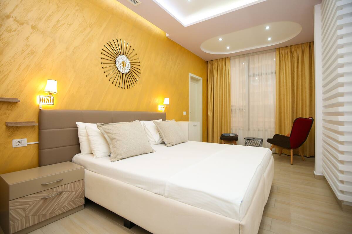 A modern hotel room with a large bed, bedside lamps, a clock on the wall, a chair, and a desk with a lamp.