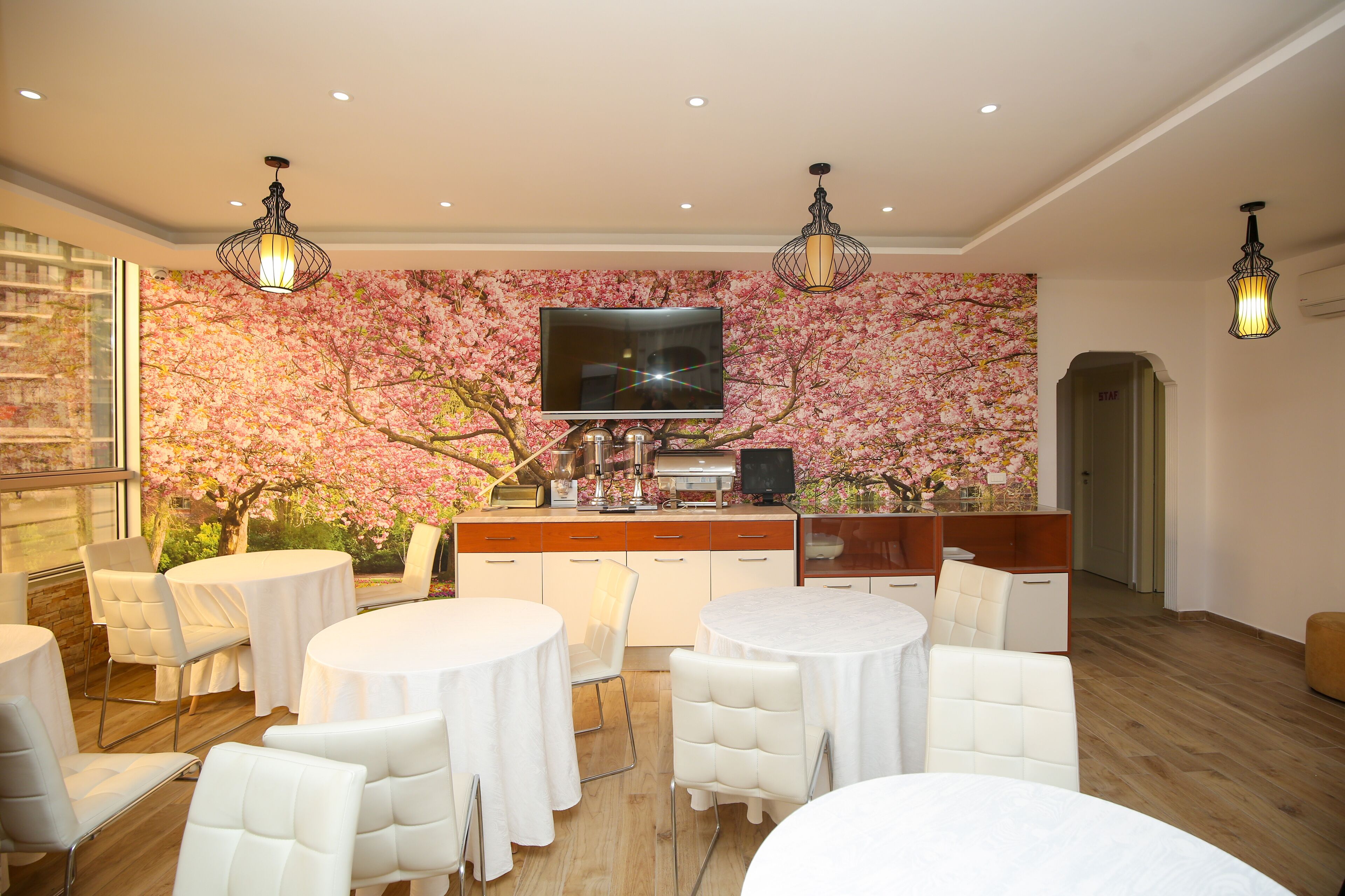 A modern dining area with round tables and white chairs, a flat-screen TV mounted on a cherry blossom wallpaper, and a wooden counter with a coffee machine.