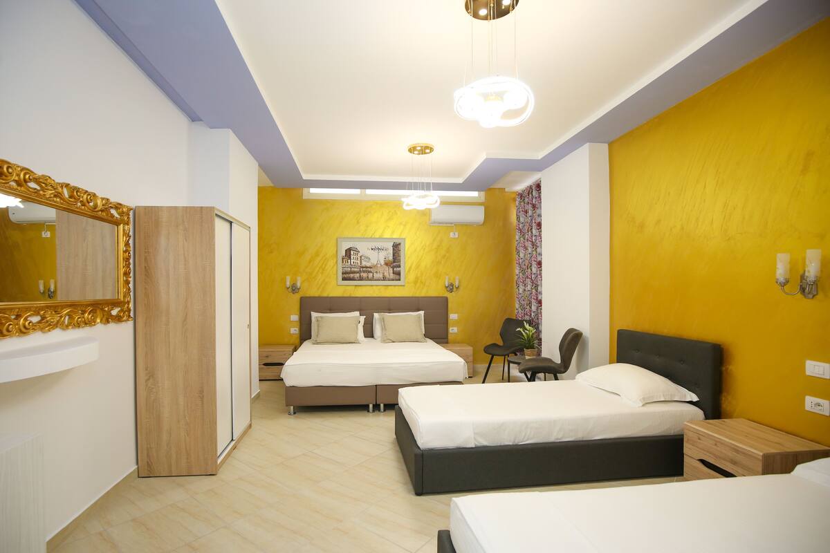 A hotel room with two beds, a large mirror, a wooden wardrobe, a headboard with a picture, and a small table.