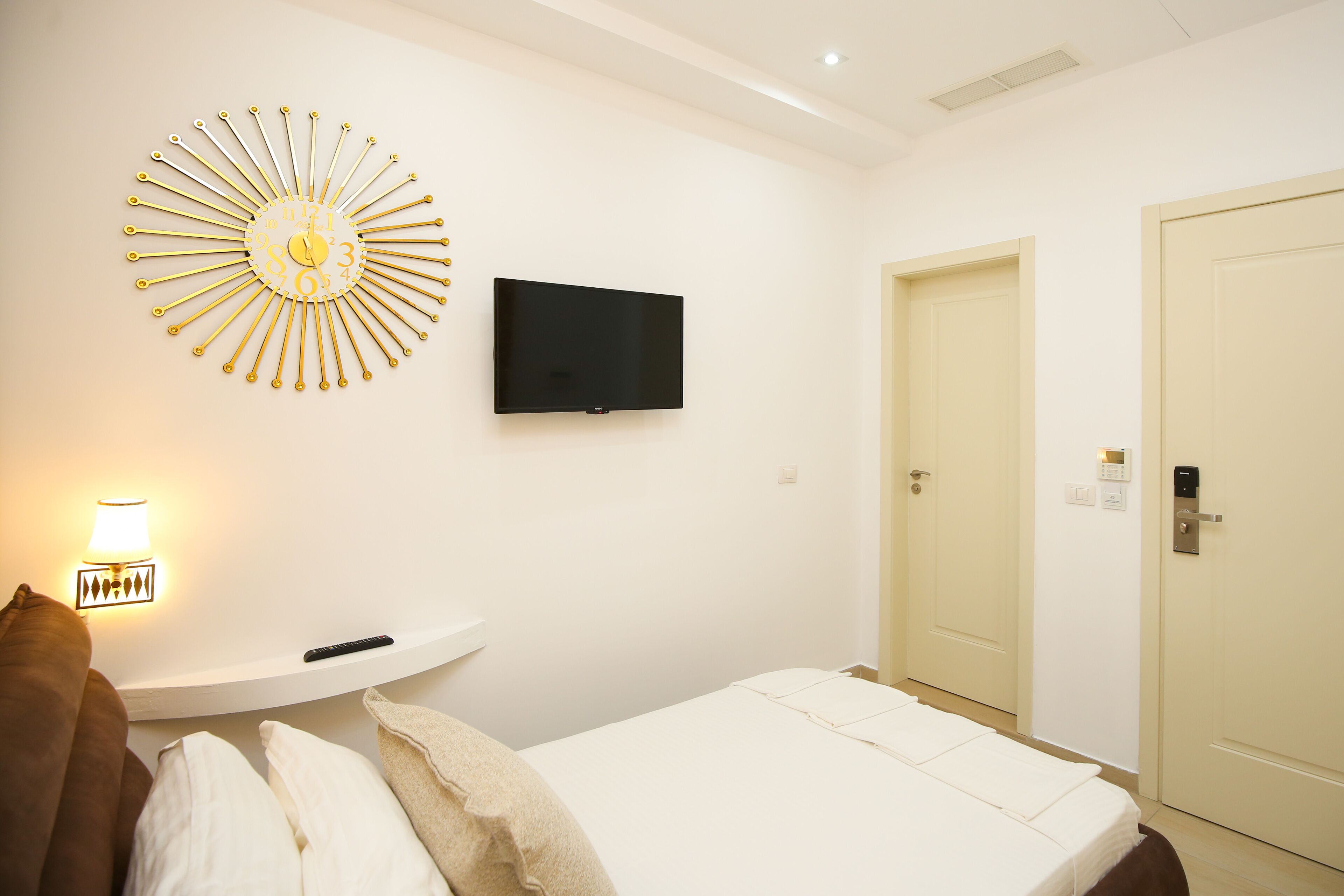 A hotel room with a bed, a wall-mounted TV, a sunburst light fixture, a bedside table with a remote control, and a door.