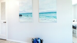 A hallway with two beach paintings on the wall, a door with louvered glass, and a pair of shoes on the floor. - Cozy 2-bed, 1-bath second-floor unit minutes from Indian Rocks Beach. Sleeps eight with full kitchen, WiFi, smart TV, beach gear, parking, and direct pool access. A comfortable, convenient stay for families and groups (Indian Rocks Beach)