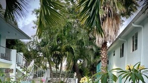 A pathway leading to a white building with a staircase and a palm tree in the center. - Cozy 2-bed, 1-bath second-floor unit minutes from Indian Rocks Beach. Sleeps eight with full kitchen, WiFi, smart TV, beach gear, parking, and direct pool access. A comfortable, convenient stay for families and groups (Indian Rocks Beach)