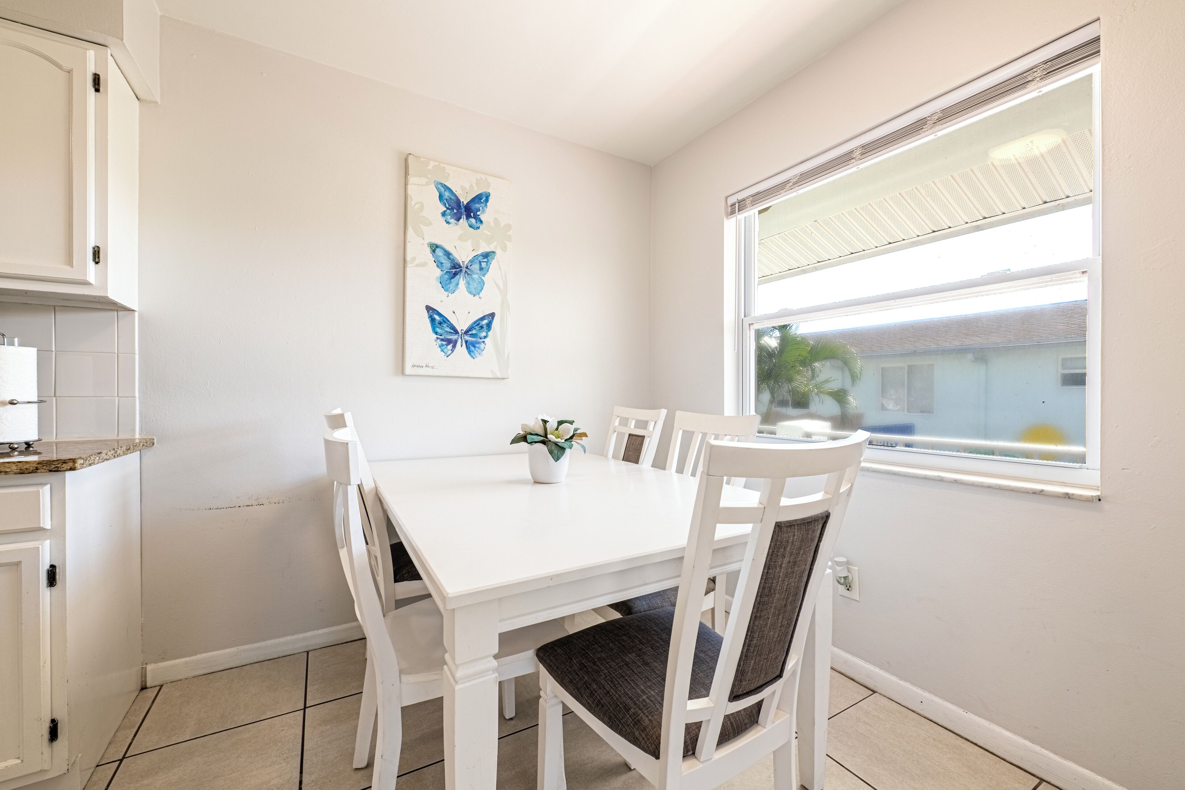 A white dining table with four chairs, a window with blinds, and a wall-mounted butterfly art piece.