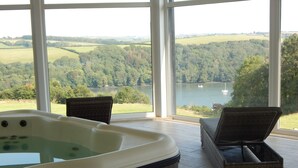 Un bain à remous sur une terrasse en bois, agrémentée de fauteuils en osier, avec vue sur un lac et des collines boisées.