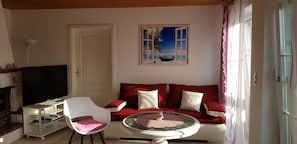 Ein Wohnzimmer mit rotem Sofa, weißem Sessel, Fernseher auf einem Schrank, einem Bild mit Strandmotiv und einer Tür, die den Blick ins Freie erlaubt.